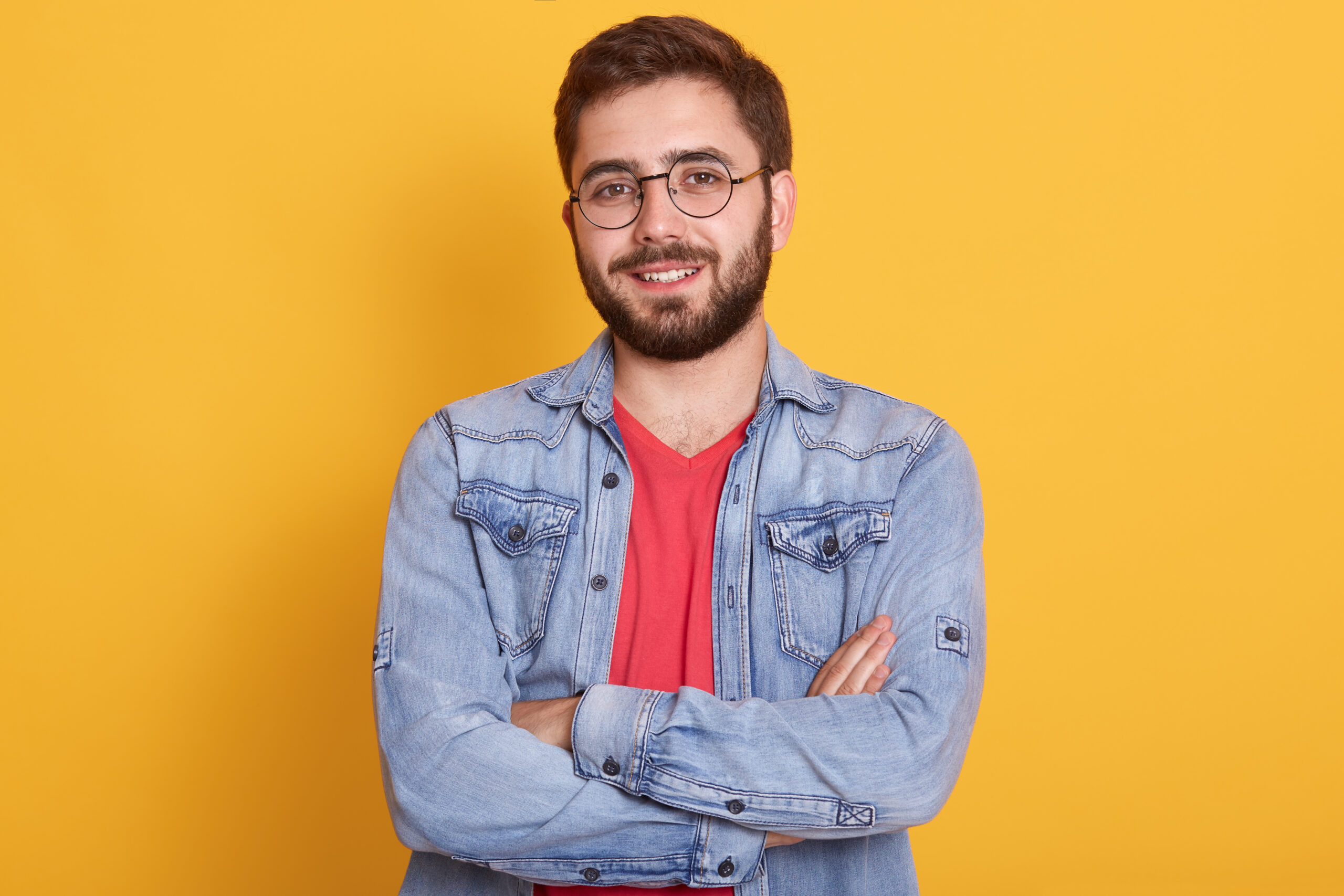 Indoor picture of cheerful handsome young man having folded hands, looking directly at camera, smiling sincerely, wearing casual clothes, posing isolated over yellow background. Emotions concept.